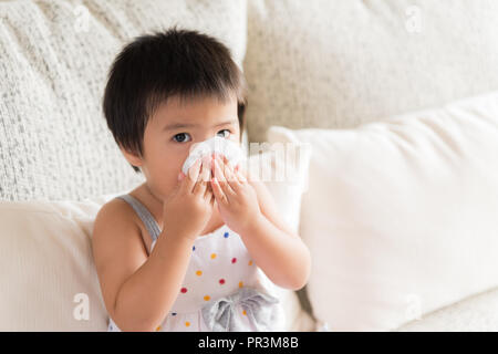 Kranken kleinen asiatischen Mädchen wischen oder Reinigung der Nase mit einem Taschentuch, auf dem Sofa zu Hause sitzen. Medizin und Gesundheit Konzept. Stockfoto