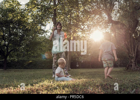 Junge Mutter bläst Seifenblasen auf ihre beiden Jungs, im Freien mit Hintergrundbeleuchtung von der schönen Sonne durch Baumkronen, getönten retro Effekt. Stockfoto