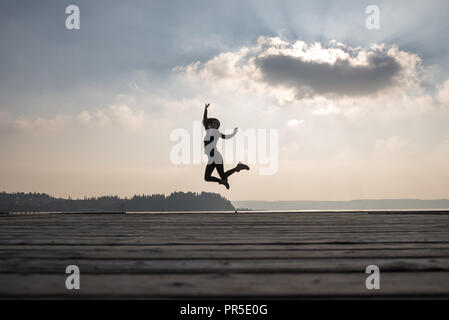 Glückliche Frau springen in die Luft vor Himmel mit Sonne hinter Wolke. Stockfoto
