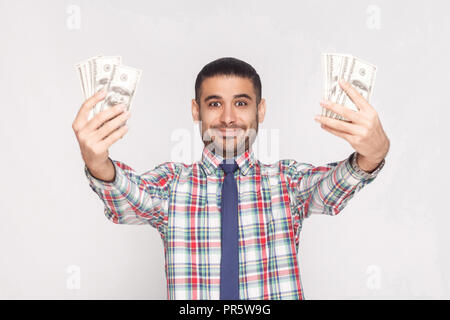 Portrait von zufriedenen Prahlende hübscher junger Geschäftsmann in kariertem Hemd mit blauer Krawatte stehen und hält das Gebläse von Dollar mit erhobenen Armen. Pflege Stockfoto