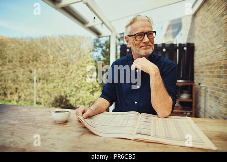 Lächelnd älterer Mann, trinken eine Tasse Kaffee und eine Zeitung lesen, während Sie sich einen Tisch draußen auf der Terrasse Stockfoto