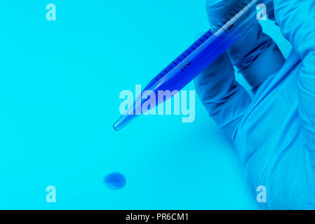 Eine behandschuhte Hand hält eine lab Pipette mit blauer Flüssigkeit auf einem blauen Hintergrund. Stockfoto