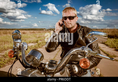 Biker sprechen auf einem Smartphone. Biker Mann mit einer Lederjacke und Sonnenbrille sitzt auf seinem Motorrad. Stockfoto