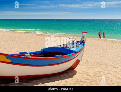 Fischerboot in traditionellen Farben gemalt, auf Armação de Pera am Strand mit einem weit entfernten Paar am Strand Stockfoto