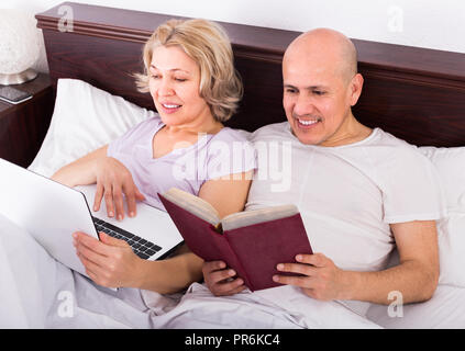 Gerne reifer Mann und Frau im Bett liegend mit Laptop und Buch und lächelnd. Fokus auf den Menschen Stockfoto