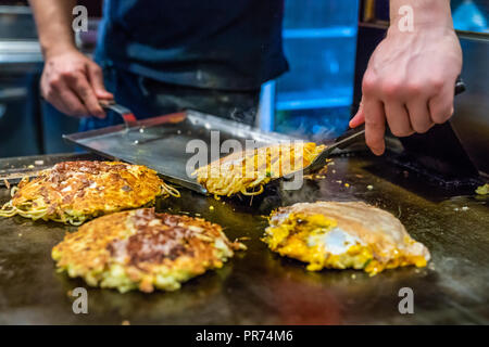 Chef kocht Okonomiyaki auf der Eisenplatte, er ist mit dem Spachtel das Essen auf die Platte, die bereits erfolgt ist, Stockfoto