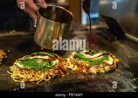 Die köstlichen okonomiyaki ist fast auf der Eisenplatte, der Koch ist Hinzufügen einige japanische traditionelle Sauce Stockfoto