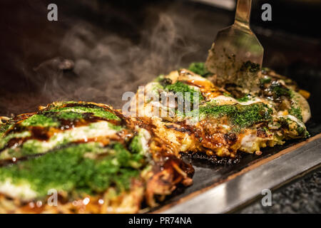 Koch mit Spachtel die getan okonomiyaki in der Hälfte auf der Eisenplatte zu schneiden, und bereit, die leckere traditionelle japanische Lebensmittel an den Kunden Stockfoto