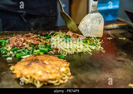 Koch ist mit dem Spachtel okonomiyaki auf der Eisenplatte zu kochen, stellt er einige Scheiben Fleisch und Gemüse in der Nahrung Stockfoto