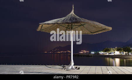 Abend noch Leben mit einem isolierten Bambus Sonnenschirm am Strand am Meer. Im Hintergrund ist die beleuchtete Stadt und das Biokovo Gebirge Stockfoto