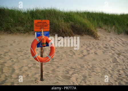 Orange Sicherheit Ring (Boje) und Rettungsschwimmer Seil hängend auf Metall Rack mit einem Metall Warnschild auf einem roten Sandstrand, Kanada Stockfoto
