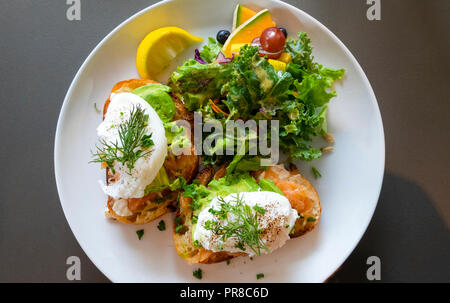 Eier Royale, pochierte Eier mit geräuchertem Lachs (Graved Lachs) und Avocado mit gemischten frisches Obst and​ Salat auf einem weißen Teller Stockfoto