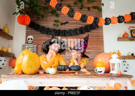 Süße kleine Mädchen und ihre Mutter, beide trugen Hexe Hüte, hinter einem Tisch in Halloween Thema eingerichtete Zimmer sitzen, einander und lächelnd. Stockfoto