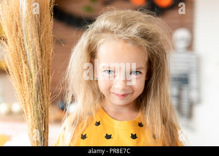 Süße kleine Mädchen mit unordentlichen Haar, verkleidet als eine Hexe und einen Besen in Halloween eingerichtetes Wohnzimmer steht, in die Kamera lächeln. Stockfoto