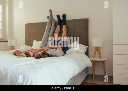 Glückliches Paar auf dem Bett zusammen liegenden Hände halten einander an. Paar liegend auf dem Bett mit den Beinen an der Wand angehoben und sprechen. Stockfoto