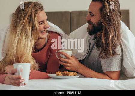 Glückliches Paar holding Kaffeetassen entspannen auf Bett mit Snacks. Lächelndes Paar liegend auf Bett in einer gemütlichen Decke sich einander, während Sie breakfa Stockfoto