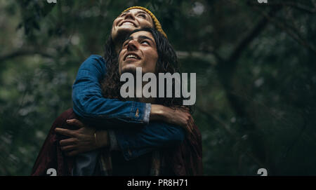 Junges Paar an Wald in Regen huckepack. Mann, der seine Frau, die auf zurück in den Wald an einem regnerischen Tag, sowohl mit der Suche und lächelnd. Stockfoto