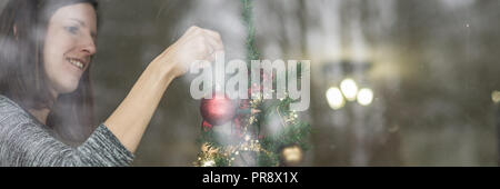 In der breiten Ansicht Bild einer jungen lächelnde Frau hängenden roten Urlaub Ball auf einen Weihnachtsbaum. Blick durch ein Fenster. Stockfoto
