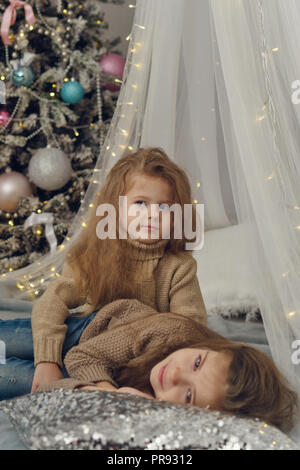 Zwei kleine Schwestern warten auf Weihnachten. Mädchen liegen auf dem Bett mit Girlande. Im Hintergrund ist Weihnachtsbaum mit Ornamenten. Sie sind in Jeans gekleidet Stockfoto