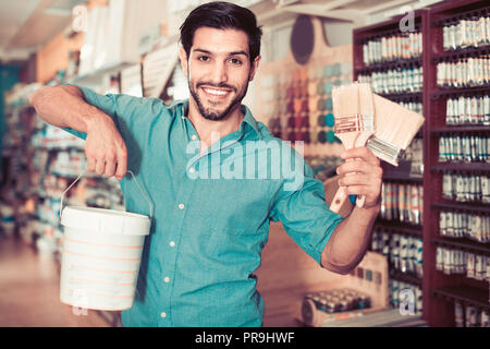 Junge positive Mann unter den Regalen in Farbe Speicher mit Pinsel und Farbe zufrieden Stockfoto