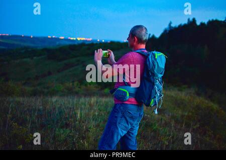 Aktiv im mittleren Alter Mann stand auf einem Hügel mit Rucksack, wobei Bild der Berge Landschaft während der Dämmerung. Mann, Bild per Handy Stockfoto