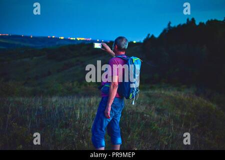 Aktiv im mittleren Alter Mann stand auf einem Hügel mit Rucksack, wobei Bild der Berge Landschaft während der Dämmerung. Mann, Bild per Handy Stockfoto
