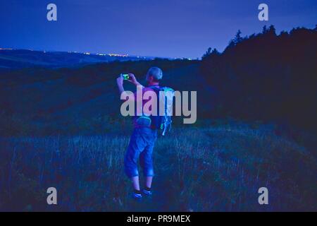 Aktiv im mittleren Alter Mann stand auf einem Hügel mit Rucksack, wobei Bild der Berge Landschaft während der Dämmerung. Mann, Bild per Handy Stockfoto