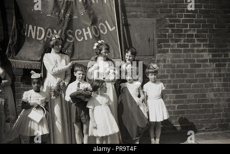 1950er Jahre, historische, Sheffield, junge Kinder und Mütter stehend außerhalb in einer Straße unter einer Fahne für die Wesleyan Methodist Church Sonntagsschule, vor der Teilnahme an einer Parade. Christlichen Charakter, eine Sonntagsschule war eine Bildungseinrichtung, zuerst in den 1780er Jahren in England gegründet, um eine Ausbildung zu Arbeiterklasse Kinder zur Verfügung zu stellen und besonders beliebt waren im nördlichen Englischen Städte wie Manchester und Sheffield. Die Lehre der Wesleyan Kirche, genannt für die Rechte der Frau und trat gegen Kinderarbeit. Stockfoto