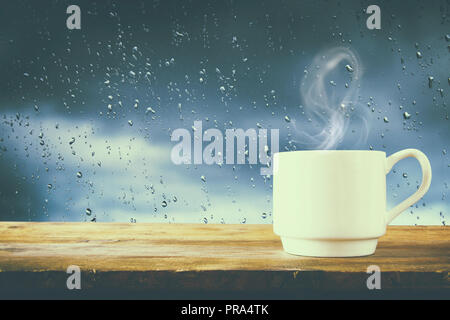 Tasse Kaffee an einem regnerischen Tag über Holz- Tisch und Fenster mit Regentropfen Hintergrund Stockfoto