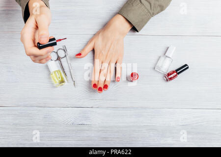 Junge Frau die Maniküre zu sich. Weibliche Hände auftragen Rot polnischen auf die Nägel neben Schere, Schaber, Flaschen mit Glitzer Lack, Base und moistur Stockfoto