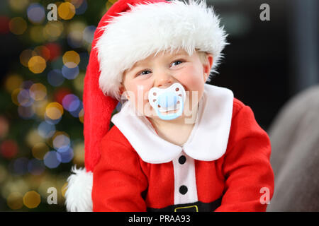 Baby Tragen santa verkleidet mit einem Schnuller im Mund an der Kamera in Weihnachten auf einer Couch im Wohnzimmer zu Hause suchen Stockfoto