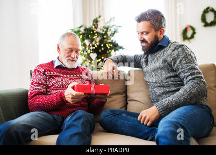 Ein älterer Vater und erwachsener Sohn mit einer gegenwärtigen Sitzen auf einem Sofa in der Weihnachtszeit. Stockfoto