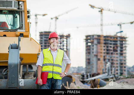 Gerne Ingenieur vor der Bagger auf einer Baustelle mit Gebäuden und Krane im Hintergrund Stockfoto