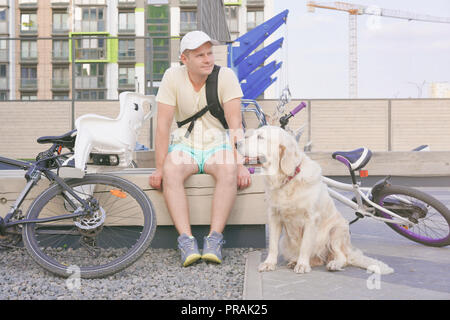 Das moderne Leben in einer großen Stadt - sportlichen jungen Mann mit Hund ruht auf einer Bank in der Stadt nach einer Radtour Stockfoto