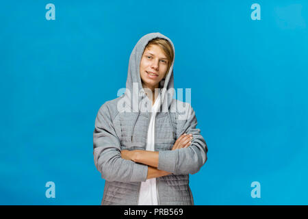 Freundlich lächelnden Teenager in grau Bluse mit Kapuze und kreuzte die Arme vor blauem Hintergrund studio Shot, isoliert Stockfoto