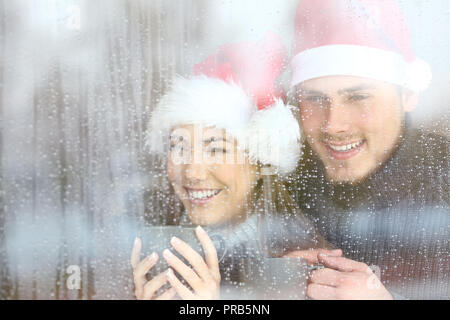 Glückliches Paar durch ein Fenster in der Weihnachtszeit in einer regnerischen Tag suchen Stockfoto