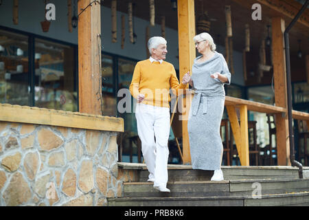 Lächelnd gut aussehender älterer Herr in Gelb pullover Unterstützung der Frau und hielt ihre Hand, während sie sich nach unten bewegenden Treppen und Chatten mit Veranda Stockfoto