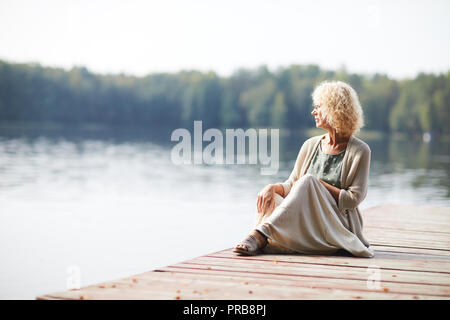 Ernste nachdenkliche lockigen Haaren reife Lady in langen Rock sitzt auf Pier und ruhigen Natur rund um Stockfoto