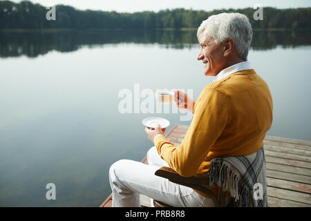 Fröhliche zierliche stattlicher Mann in gelb Pullover und weißen Hosen sitzen auf Stuhl mit Decke auf dem Pier und trinken lecker Kaffee, während der Betrachtung Stockfoto