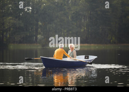 Lächelnd schönen lockigen Haaren ältere Dame in Cardigan sitzen auf dem Boot und im Gespräch mit Freund, während er Rudern Rudern Stockfoto