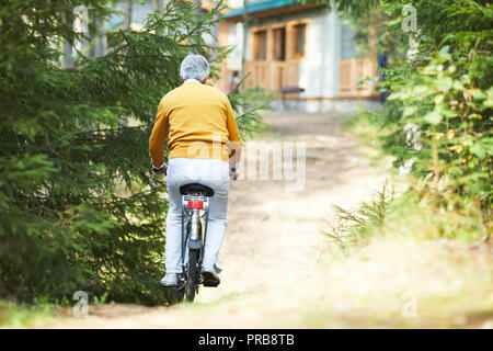 Ansicht der Rückseite des Grauhaariger alter Mann in Gelb pullover Reiten Fahrrad bis zu Country House, Aktive ältere Menschen Radfahren auf rauem Weg Stockfoto