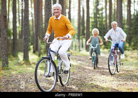 Fröhlich aufgeregt senior Freunden in Freizeitkleidung genießen Leben aktiv Radfahren auf dem Fahrrad zusammen im Wald, sie rennen sich gegenseitig Stockfoto