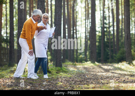 Ernst Ärztin in Laborkittel holding Zwischenablage und die Unterstützung älterer Patienten während im Wald spazieren, Wiederherstellungsverfahren Stockfoto