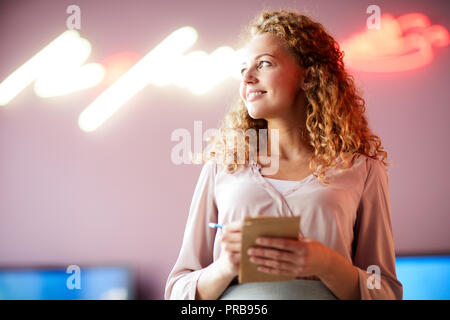 Lächelnd zielgerichtete schöne Mädchen mit lockigem Haar holding Notizblock und Stift und Wegsehen, während im Cafe arbeiten mit neon Inschrift an der Wand Stockfoto