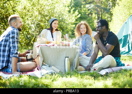 Vier fröhliche interkulturelle Freunde sitzen um selbstgebaute Tabelle in der grünen Wiese, in Reden und Essen Stockfoto