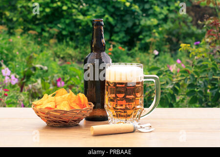 Glas Krug Bier und eine Flasche Bier auf Holztisch mit Kartoffelchips in Weidenkorb und Öffner auf natürlichen, grünen Hintergrund Stockfoto