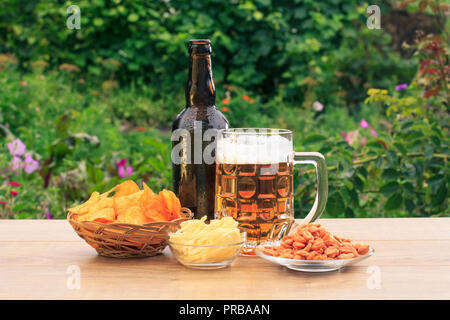 Glas Krug Bier und eine Flasche Bier auf Holztisch mit Kartoffelchips in Weidenkorb, Erdnüsse und getrockneten Tintenfisch in Schalen auf natürlichen, grünen Hintergrund Stockfoto