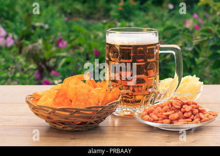 Glas Becher Bier auf Holztisch mit Kartoffelchips in Weidenkorb, Erdnüsse und getrockneten Tintenfisch in Schalen auf natürlichen, grünen Hintergrund Stockfoto