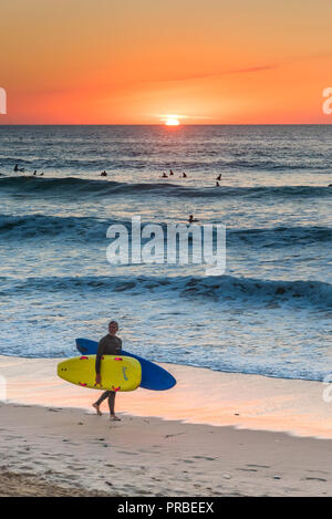 Ein Surfer die Surfbretter entlang der Küste wie die Sonne auf den Fistral Beach in Newquay in Cornwall. Stockfoto