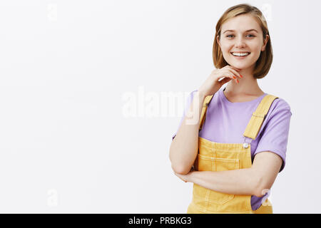 Mädchen feste Laptop von sich selbst, das Gefühl Stolz und Ruhm. Gerne attraktive blonde Frau mit kurzen modischen Haarschnitt, stehend in Overalls und starrte mit intrigiert Lächeln an Kamera, Kinn berühren. Stockfoto